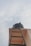 Looking up towards a black water tank sitting atop a building alongside a dark metal ladder leading up to it. The scene is set against a backdrop of a cloudy sky.