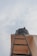 Looking up towards a black water tank sitting atop a building alongside a dark metal ladder leading up to it. The scene is set against a backdrop of a cloudy sky.