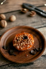 A rustic wooden table holds a round wooden plate with a baked dessert topped with nuts and a caramel-like glaze. Surrounding the dessert are several pieces of dark chocolate. In the background, some walnuts and a nutcracker are visible.