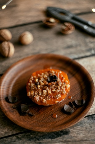 A rustic wooden table holds a round wooden plate with a baked dessert topped with nuts and a caramel-like glaze. Surrounding the dessert are several pieces of dark chocolate. In the background, some walnuts and a nutcracker are visible.