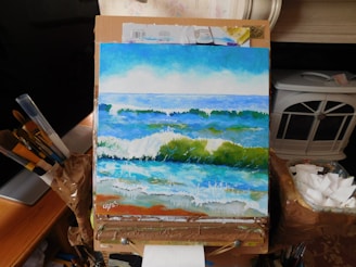 A painting of an ocean scene with waves crashing onto the shore, displayed on an easel surrounded by art supplies like paintbrushes and tubes of paint. The painting captures the movement and energy of the sea with rich blues and greens.