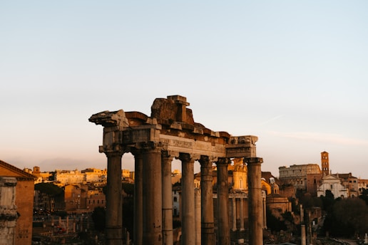 Ancient Roman ruins with tall stone columns stand prominently under a clear sky with warm lighting giving a golden hue to the structures. Surrounding the ruins are historical buildings in a city landscape.