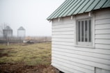 A misty farm scene with a white wooden shed featuring a textured window and a green-tinted roof. Two silos are partially visible in the background, shrouded in fog. The ground is a mix of grass and bare earth, adding to the rural setting.