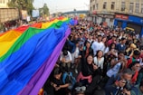 A large group of people participating in a street parade, carrying a vibrant rainbow flag, suggestively for a pride event. The crowd appears diverse and cheerful, with individuals displaying expressions of joy and excitement. The surroundings include urban buildings and a street filled with signs and banners.