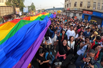 A large group of people participating in a street parade, carrying a vibrant rainbow flag, suggestively for a pride event. The crowd appears diverse and cheerful, with individuals displaying expressions of joy and excitement. The surroundings include urban buildings and a street filled with signs and banners.