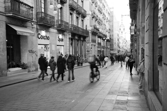 A bustling urban street scene in black and white, showing people walking and cycling along a cobblestone street lined with historic buildings. The architecture features balconies and street lamps, and there are various shops and businesses with signs such as 'Corcha' and 'Spain'. The street is lively, with individuals engaged in different activities, suggesting a dynamic city atmosphere.