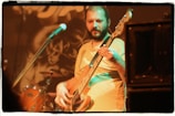 A musician plays an electric bass guitar on stage, surrounded by musical equipment. The background features a blurred image of drums and a microphone. The warm lighting creates a focus on the musician's expression as he concentrates on playing.