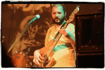 A musician plays an electric bass guitar on stage, surrounded by musical equipment. The background features a blurred image of drums and a microphone. The warm lighting creates a focus on the musician's expression as he concentrates on playing.
