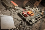 A setup of various guitar effects pedals arranged on a pedalboard, surrounded by numerous cables. The pedals include various colors such as red, orange, and grey, indicating different types of sound effects. A paper with a setlist or notes is partially visible on the floor.