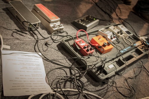 A setup of various guitar effects pedals arranged on a pedalboard, surrounded by numerous cables. The pedals include various colors such as red, orange, and grey, indicating different types of sound effects. A paper with a setlist or notes is partially visible on the floor.