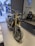 A Triumph motorcycle is prominently displayed indoors with a showroom setting. The bike has a sleek design with gold front forks and black detailing. In the background, there is an image of motorcyclists riding on a road. The showroom features a Triumph logo and the phrase 'For The Ride' on the wall, along with an office chair and desk nearby.