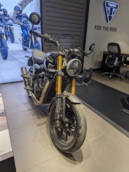 A Triumph motorcycle is prominently displayed indoors with a showroom setting. The bike has a sleek design with gold front forks and black detailing. In the background, there is an image of motorcyclists riding on a road. The showroom features a Triumph logo and the phrase 'For The Ride' on the wall, along with an office chair and desk nearby.