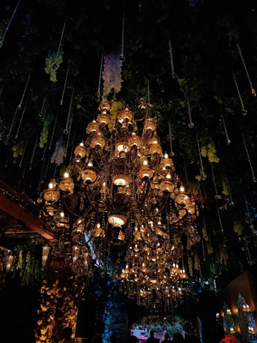 A grand and ornate chandelier hangs from the ceiling, illuminated with numerous warm lights. The setting appears to be a lavish indoor space, adorned with hanging plants and floral decorations. The light from the chandelier casts a rich glow, creating an opulent ambiance.