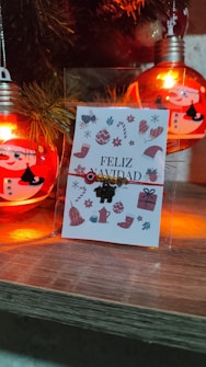 A festive scene featuring Christmas decorations, including a card with the text 'Feliz Navidad' and various holiday-themed illustrations such as mittens, hats, and presents. The card is positioned next to decorative light bulbs painted with snowman faces and surrounded by pine branch elements. The warm glow of the lights gives a cozy atmosphere to the wooden surface where the items are placed.