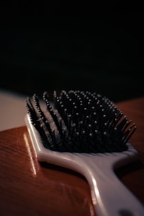 A close-up view of a hairbrush lying on a wooden surface, with focus on the bristles that have small round tips. The lighting is dim, casting shadows and creating a moody atmosphere.