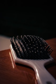 A close-up view of a hairbrush lying on a wooden surface, with focus on the bristles that have small round tips. The lighting is dim, casting shadows and creating a moody atmosphere.