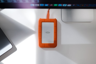 A rugged external hard drive with an orange rubberized protective casing is placed on a white desk. It is connected to a computer or device via an orange cable. The desk also features a laptop and a monitor, creating a neat workspace environment.
