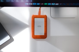 A rugged external hard drive with an orange rubberized protective casing is placed on a white desk. It is connected to a computer or device via an orange cable. The desk also features a laptop and a monitor, creating a neat workspace environment.