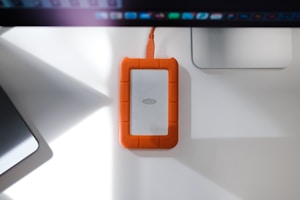 A rugged external hard drive with an orange rubberized protective casing is placed on a white desk. It is connected to a computer or device via an orange cable. The desk also features a laptop and a monitor, creating a neat workspace environment.