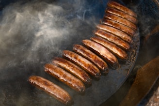 Several sausages are cooking in a pan, surrounded by steam. The sausages are positioned in a neat row and appear slightly browned and glistening from the heat. The steam adds a sense of warmth and cooking in progress.