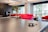 A ping pong paddle rests on top of a table tennis table. The background features a modern interior space with a large red couch, potted plants, and large windows allowing natural light.
