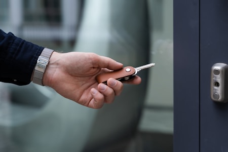 A hand is holding a set of keys with a leather keychain near a door lock. The person is wearing a silver watch and a dark button-up shirt. The background is slightly blurred, giving focus to the hand and keys.