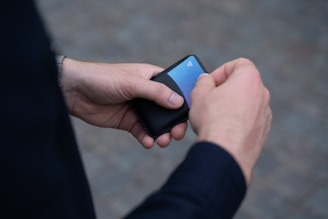 A person holding a small, black wallet with a blue credit card partially visible. The hands appear to be male, and the person is wearing a dark jacket. The watch is visible on the wrist of the left hand. The background is a blurred cobblestone surface.