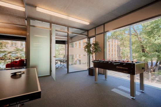 A modern office space featuring large glass windows with a view of trees and buildings outside. Inside, there is a table football game, a paddle with balls on a table, and a plant. The area appears well-lit, with a mix of natural and artificial lighting.