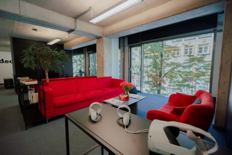 The image depicts a modern office space with a large window that offers a view of the outside greenery and a building. The office features vibrant red couches arranged around a coffee table, on which a pot of red flowers sits. A tall plant is located next to the couches. On a nearby table, a virtual reality headset and headphones are placed, adding a tech-savvy feel to the space.