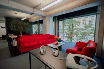 The image depicts a modern office space with a large window that offers a view of the outside greenery and a building. The office features vibrant red couches arranged around a coffee table, on which a pot of red flowers sits. A tall plant is located next to the couches. On a nearby table, a virtual reality headset and headphones are placed, adding a tech-savvy feel to the space.