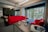 The image depicts a modern office space with a large window that offers a view of the outside greenery and a building. The office features vibrant red couches arranged around a coffee table, on which a pot of red flowers sits. A tall plant is located next to the couches. On a nearby table, a virtual reality headset and headphones are placed, adding a tech-savvy feel to the space.