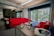 The image depicts a modern office space with a large window that offers a view of the outside greenery and a building. The office features vibrant red couches arranged around a coffee table, on which a pot of red flowers sits. A tall plant is located next to the couches. On a nearby table, a virtual reality headset and headphones are placed, adding a tech-savvy feel to the space.