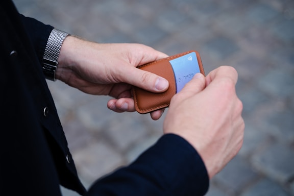 A person holding a tan leather card holder with a blue credit card partially exposed, against a blurred cobblestone background. The person's hands are prominent, and they are wearing a watch with a metal band on the left wrist. The outfit appears to include a dark long-sleeved garment.