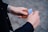 A person holding a tan leather card holder with a blue credit card partially exposed, against a blurred cobblestone background. The person's hands are prominent, and they are wearing a watch with a metal band on the left wrist. The outfit appears to include a dark long-sleeved garment.