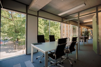 An office room with large floor-to-ceiling windows allowing plenty of natural light. Inside, there is a white rectangular table surrounded by four black office chairs. Outside, the view includes trees and a building.