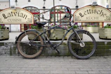 A bicycle is parked in front of a restaurant with signs advertising Latvian cuisine. The bike is placed against a brick platform, surrounded by planters filled with flowers. The background features large windows with red frames and traditional architectural details, creating a quaint and charming atmosphere.