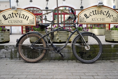 A bicycle is parked in front of a restaurant with signs advertising Latvian cuisine. The bike is placed against a brick platform, surrounded by planters filled with flowers. The background features large windows with red frames and traditional architectural details, creating a quaint and charming atmosphere.