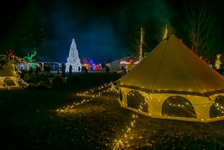 A festive scene with illuminated tents and trees adorned with colorful lights, creating a vibrant and cheerful atmosphere. A large, intricately decorated Christmas tree stands prominently in the background, surrounded by groups of people. The area is lit by various string lights, enhancing the night-time setting.