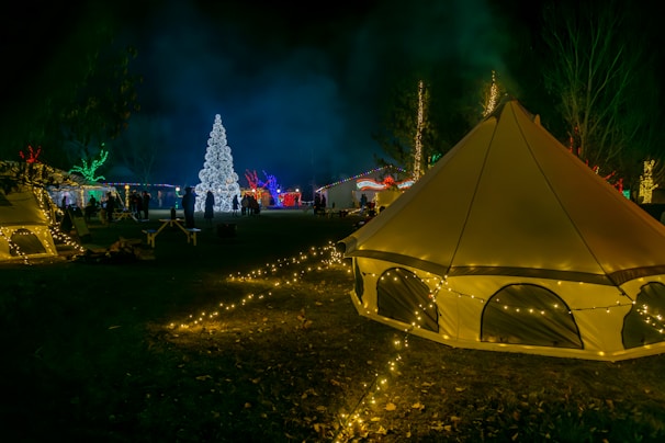 A festive scene with illuminated tents and trees adorned with colorful lights, creating a vibrant and cheerful atmosphere. A large, intricately decorated Christmas tree stands prominently in the background, surrounded by groups of people. The area is lit by various string lights, enhancing the night-time setting.
