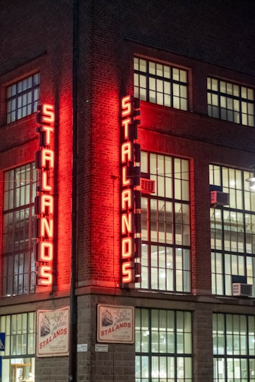 A corner of a brick building featuring tall, narrow windows adorned with a neon sign that reads 'Stalands'. The sign is illuminated in a bright red hue, casting a warm glow on the facade. The building has a classic industrial look, with visible interior lighting.