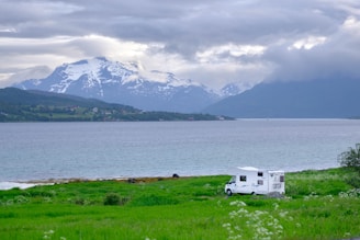 A scenic landscape featuring a large body of water with a backdrop of majestic snow-capped mountains under a cloudy sky. In the foreground, a white campervan is parked on a lush green field beside the shore. The mountains and water create a tranquil and expansive feeling.
