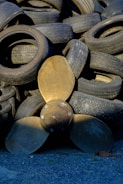 A collection of used rubber tires is stacked haphazardly, forming an uneven pile. In front of the tires, a large bronze-colored propeller with three blades is positioned upright, contrasting with the darker tones of the tires. The ground is covered with small gray gravel.