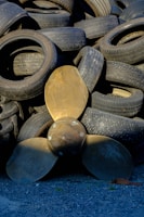 A collection of used rubber tires is stacked haphazardly, forming an uneven pile. In front of the tires, a large bronze-colored propeller with three blades is positioned upright, contrasting with the darker tones of the tires. The ground is covered with small gray gravel.