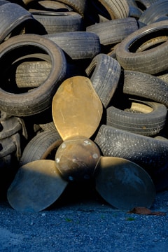 A collection of used rubber tires is stacked haphazardly, forming an uneven pile. In front of the tires, a large bronze-colored propeller with three blades is positioned upright, contrasting with the darker tones of the tires. The ground is covered with small gray gravel.