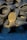 A collection of used rubber tires is stacked haphazardly, forming an uneven pile. In front of the tires, a large bronze-colored propeller with three blades is positioned upright, contrasting with the darker tones of the tires. The ground is covered with small gray gravel.