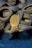 A collection of used rubber tires is stacked haphazardly, forming an uneven pile. In front of the tires, a large bronze-colored propeller with three blades is positioned upright, contrasting with the darker tones of the tires. The ground is covered with small gray gravel.