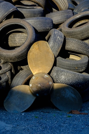 A collection of used rubber tires is stacked haphazardly, forming an uneven pile. In front of the tires, a large bronze-colored propeller with three blades is positioned upright, contrasting with the darker tones of the tires. The ground is covered with small gray gravel.