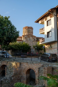 A historic stone building with an ancient architectural design stands behind a modern residential building. There's a lush green tree and several parked cars along the street. In the foreground, an old brick and stone structure is visible, possibly part of the historic scenery.