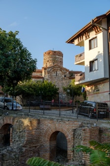 A historic stone building with an ancient architectural design stands behind a modern residential building. There's a lush green tree and several parked cars along the street. In the foreground, an old brick and stone structure is visible, possibly part of the historic scenery.