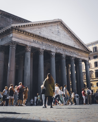 A large historical building with tall columns and an inscription on the front. Many people are standing and walking around the structure, suggesting it is a popular tourist attraction. The architecture appears to be ancient and well-preserved.